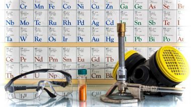 periodic table poster behind scientific tools: bunsen burner, goggles, vial of solution, gas mask.